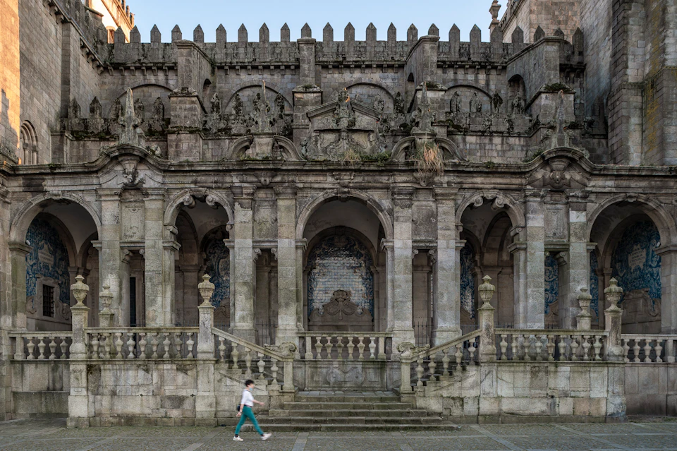 A person walks in front of an ornate, old stone building with arched entryways and detailed architectural carvings.