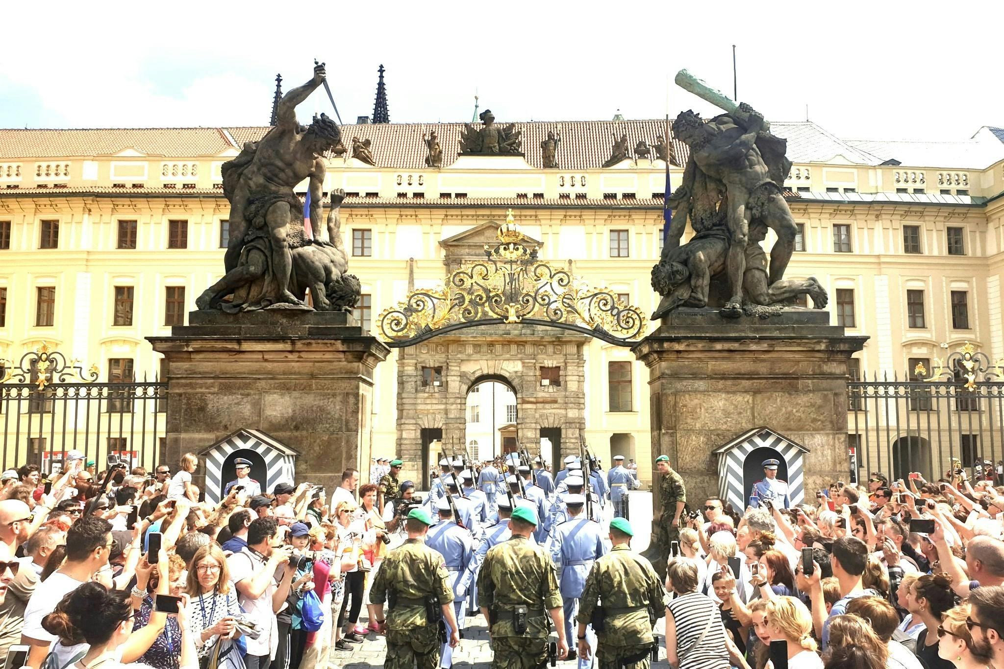 A ceremonial parade with soldiers marching through an ornate gate, flanked by large statues, as a crowd watches and takes photos.