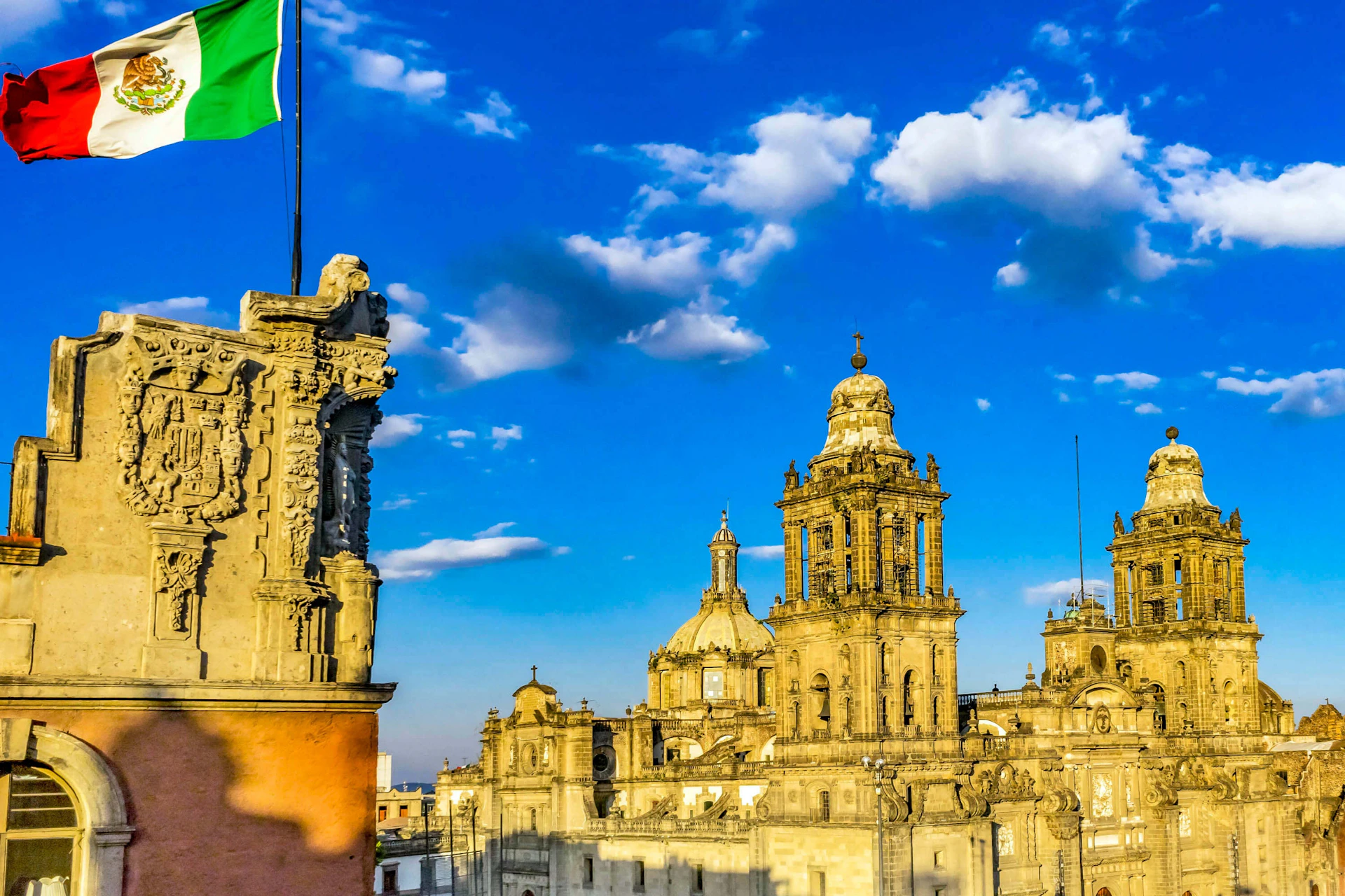 Historic cathedral with intricate architecture under a vibrant blue sky with scattered clouds. A Mexican flag is partially visible.