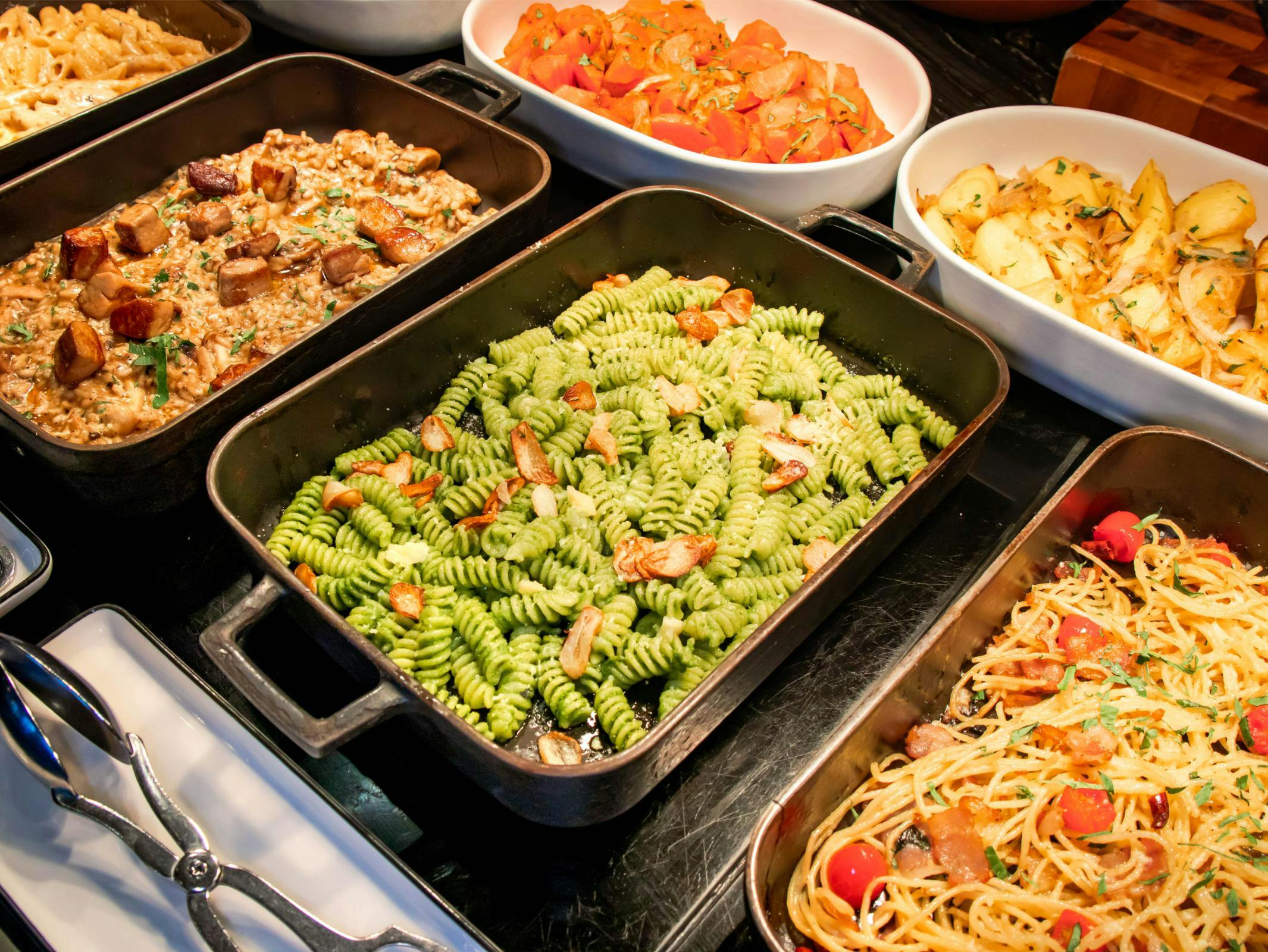 A buffet display featuring dishes such as green pasta, spaghetti, roasted vegetables, and other cooked meals in trays.