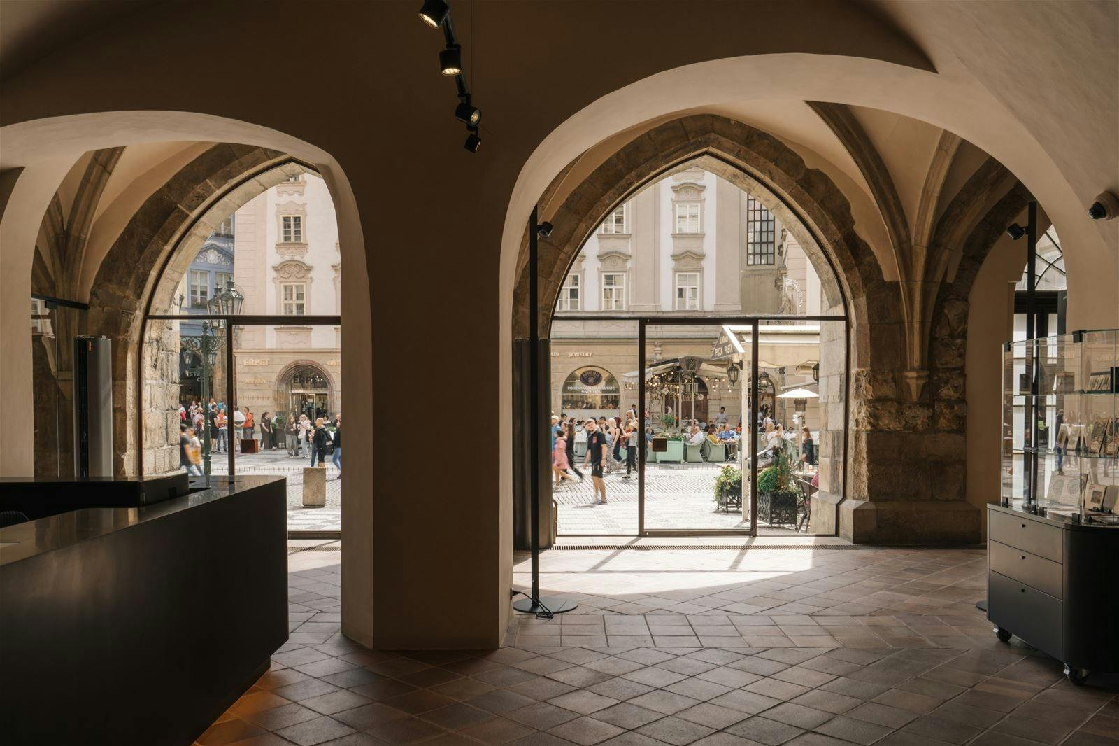 Large arched windows and doorways of a historic building overlook a busy street with people walking and old buildings in the background.