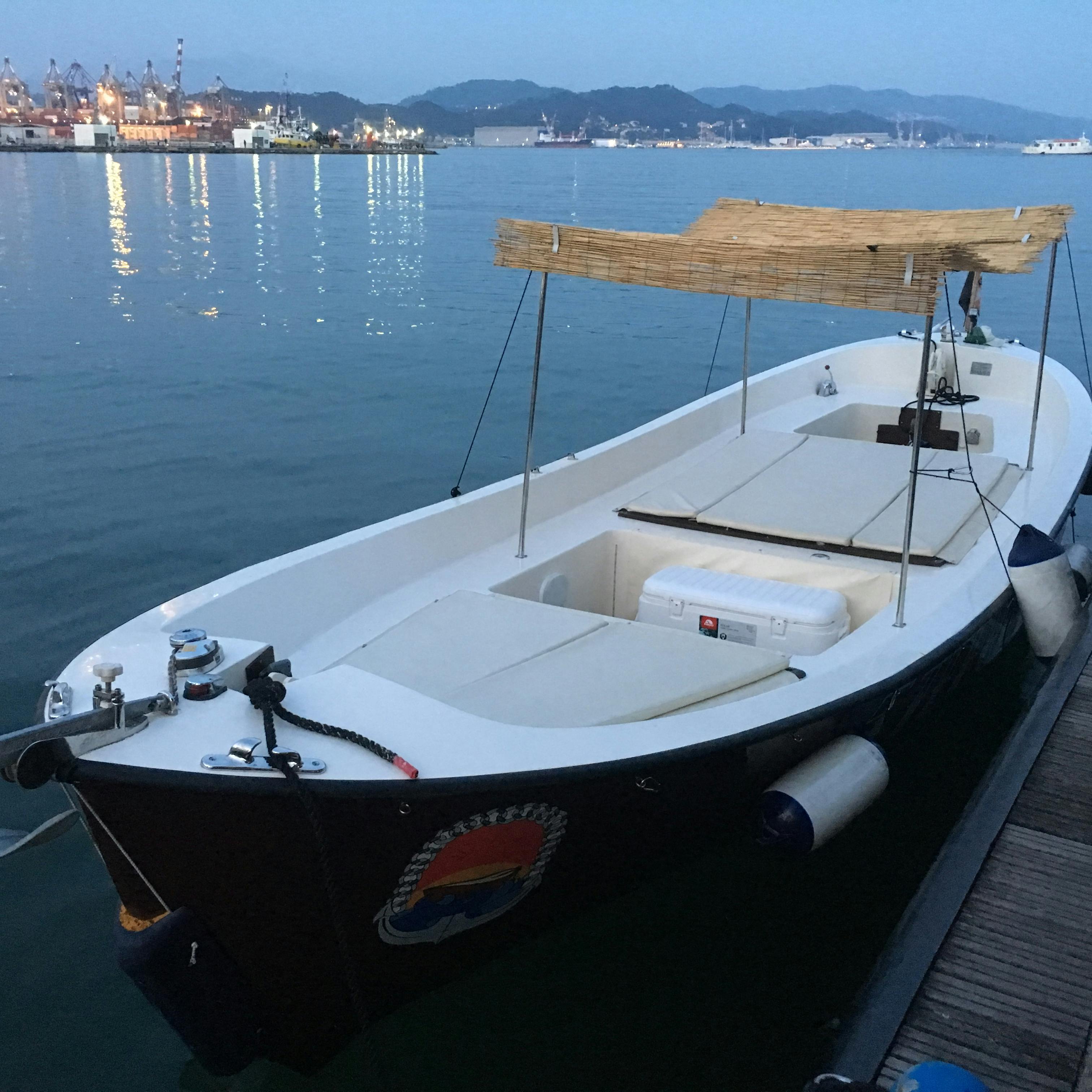 A small motorboat with a canopy is docked at a pier in a harbor during early morning or evening, with distant mountains and industrial buildings.