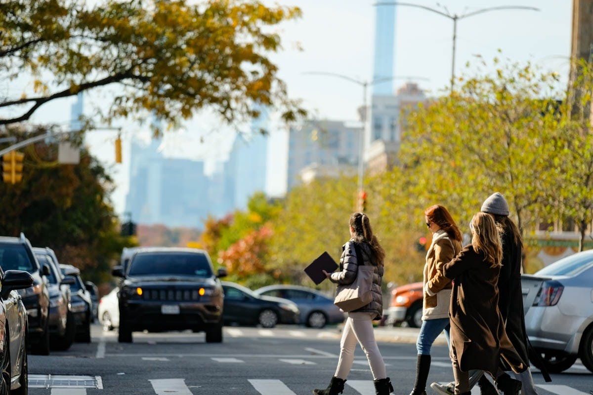 Люди переходят улицу города на пешеходном переходе, с деревьями и припаркованными автомобилями на заднем плане.