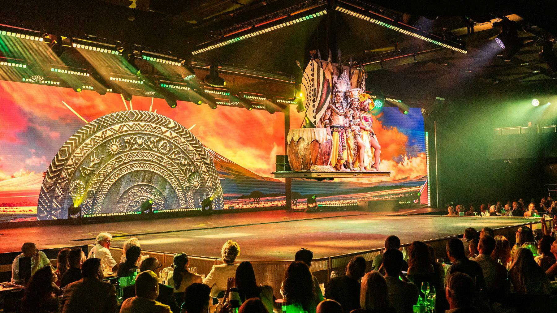 Representação em palco com um cenário colorido de temática antiga e artistas em trajes elaborados. Público em primeiro plano.