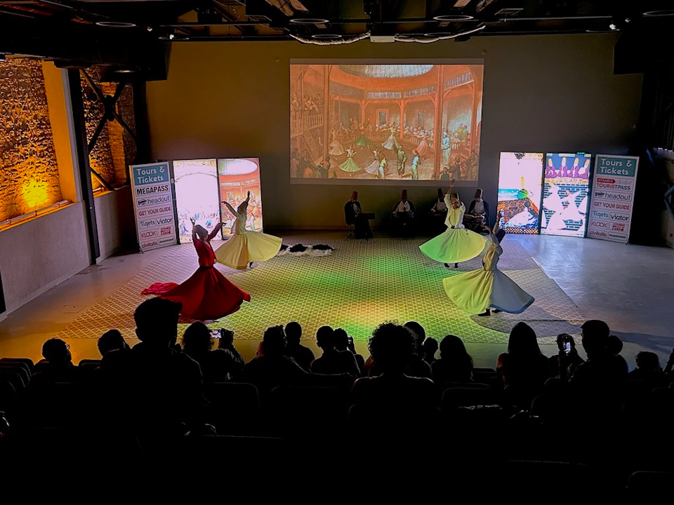 Performers in colorful dresses dance on a lit stage, watched by an audience. A historic scene is projected on the back wall.
