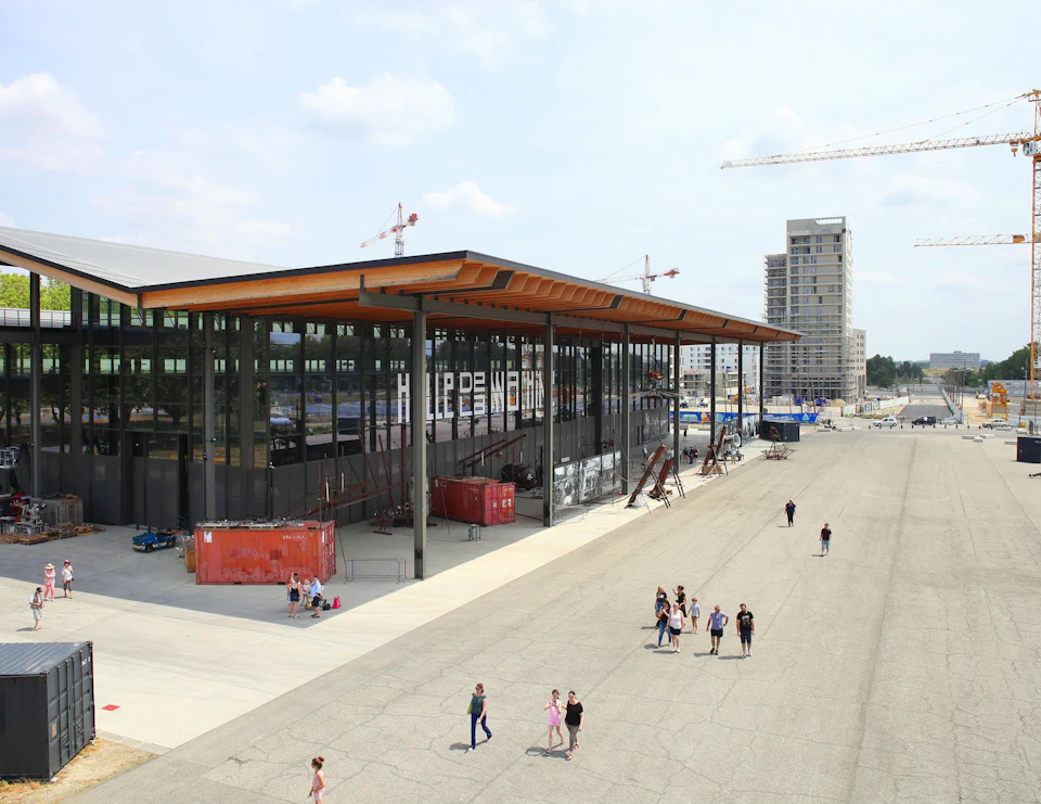 People walk on a wide pavement next to a modern, glass-covered building with cranes and a high-rise in the background.