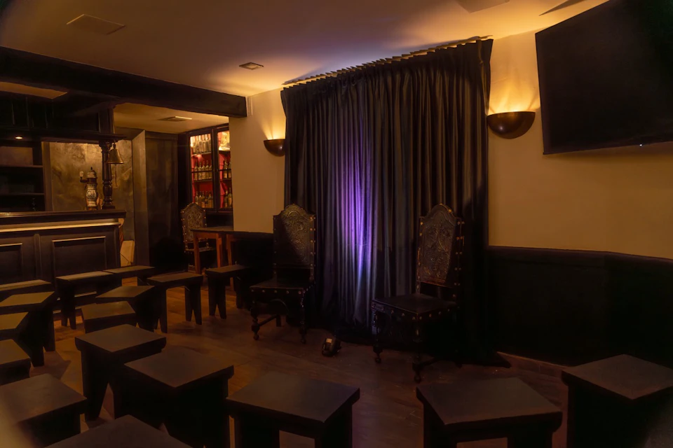Dimly lit room with two ornate chairs in front of closed curtains, and several small stools and tables arranged in rows.