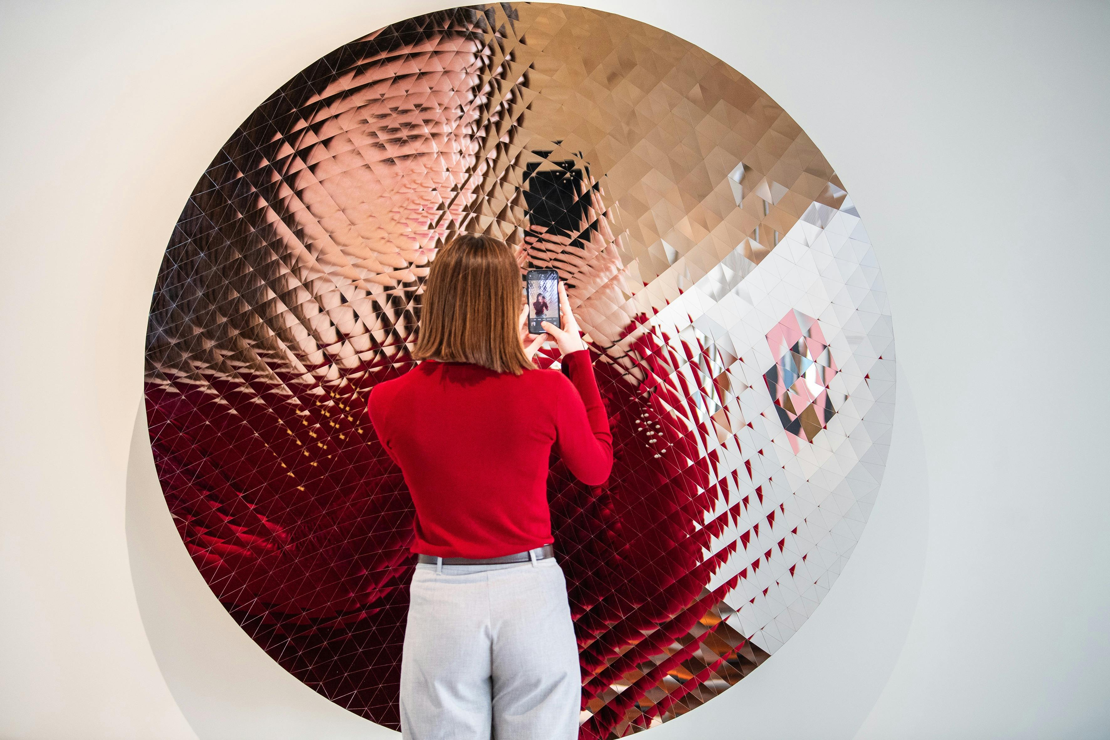 Osoba w czerwonym swetrze fotografuje swoje odbicie w dużym, teksturowanym, okrągłym lustrze z geometrycznym wzorem.