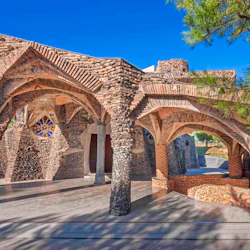 En stenbåge med invecklat tegel och glasmålningar under en klarblå himmel, med träd i bakgrunden.