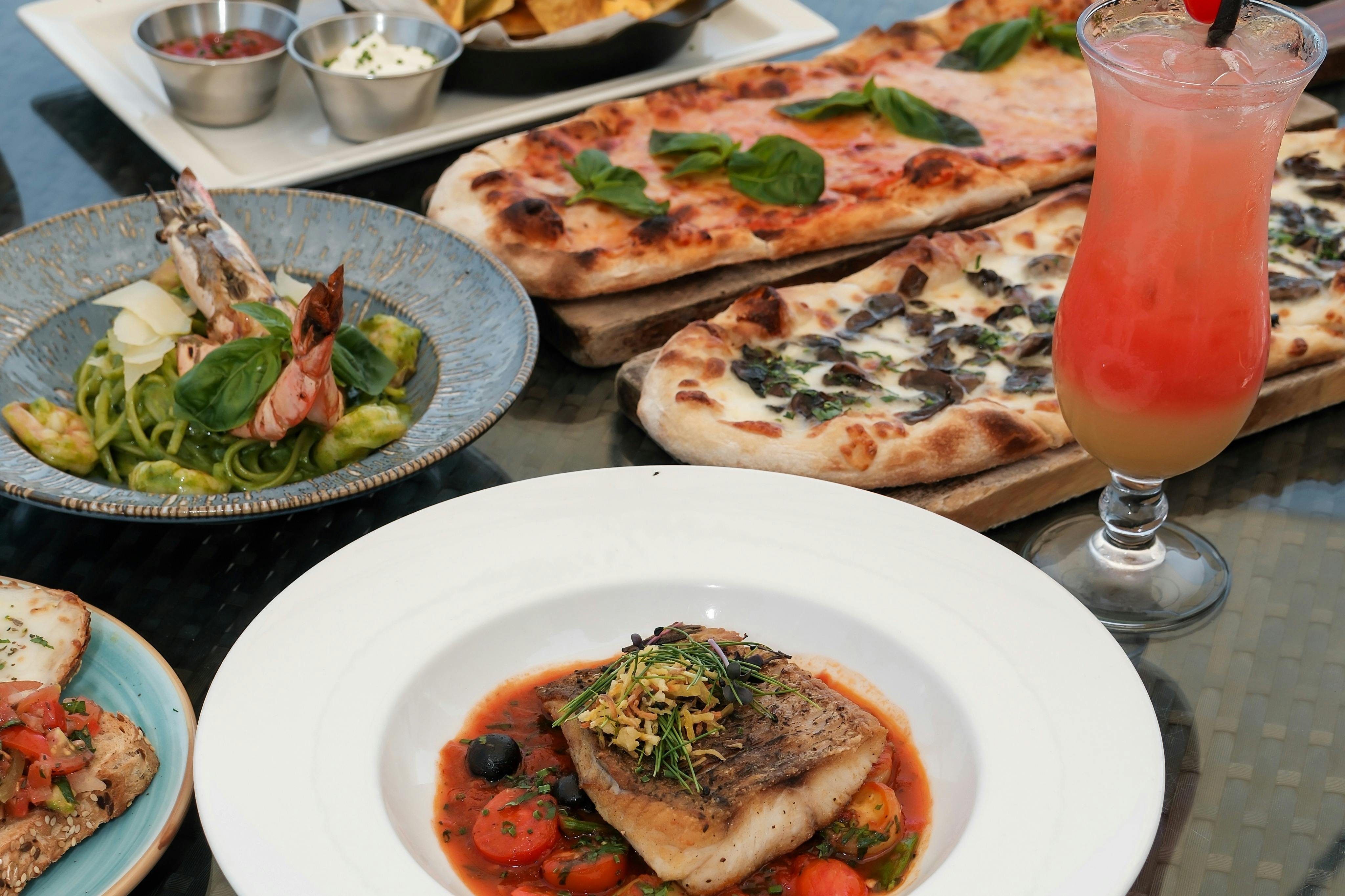 ピザ、魚の主菜、エビのパスタ、ディップのナチョス、フルーティーなカクテルなど、さまざまな料理が並ぶテーブル。