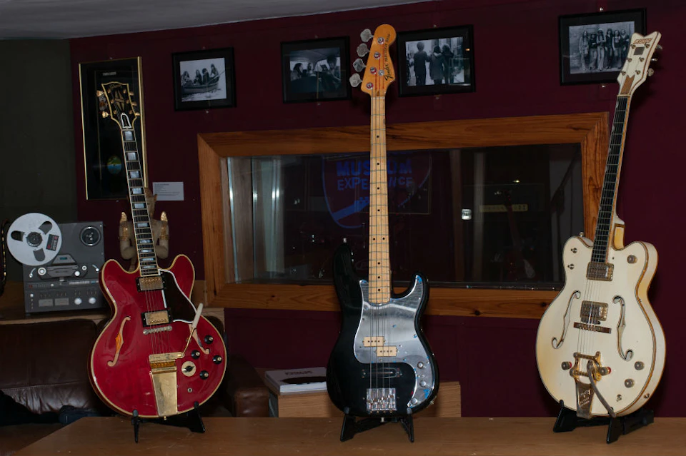 Three electric guitars on stands in a room, with framed photos on a maroon wall and a glass window in the background.