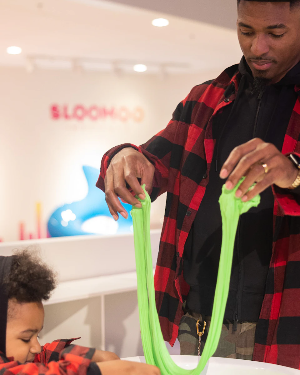 Two people in matching red plaid shirts play with green slime at a table in a brightly lit indoor space.