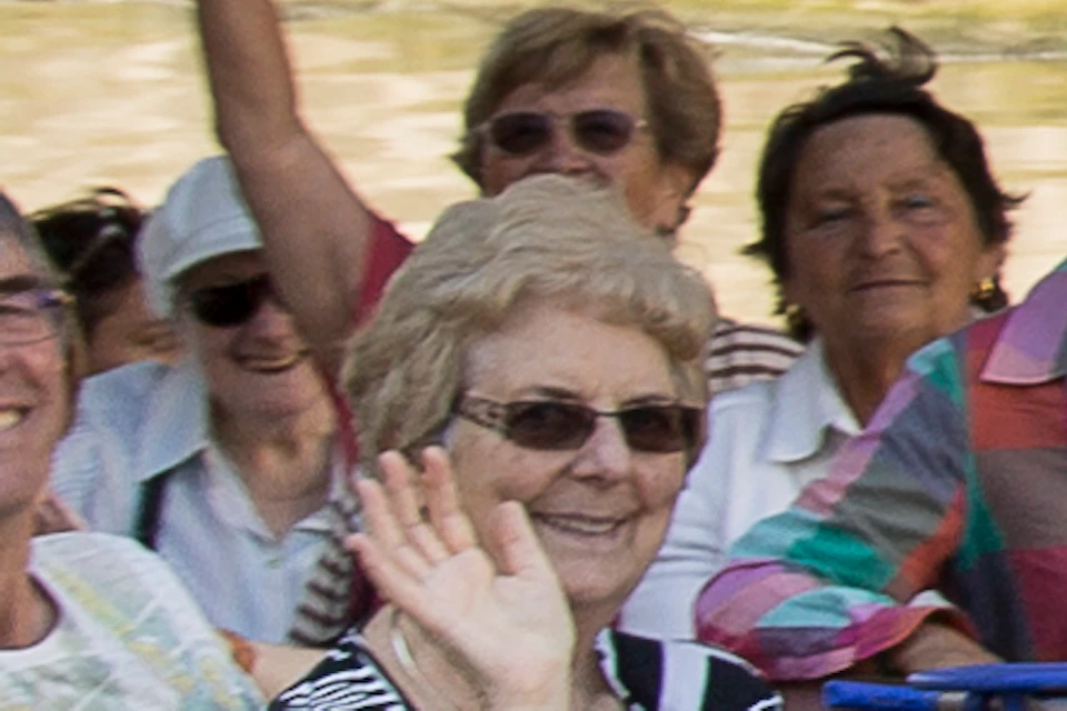 A group of people wave and smile while riding a white boat under a stone bridge on a sunny day.