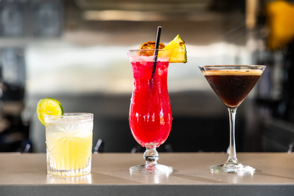 Three cocktails on a counter: a yellow drink with lime, a red drink with pineapple garnish, and a dark drink in a martini glass.