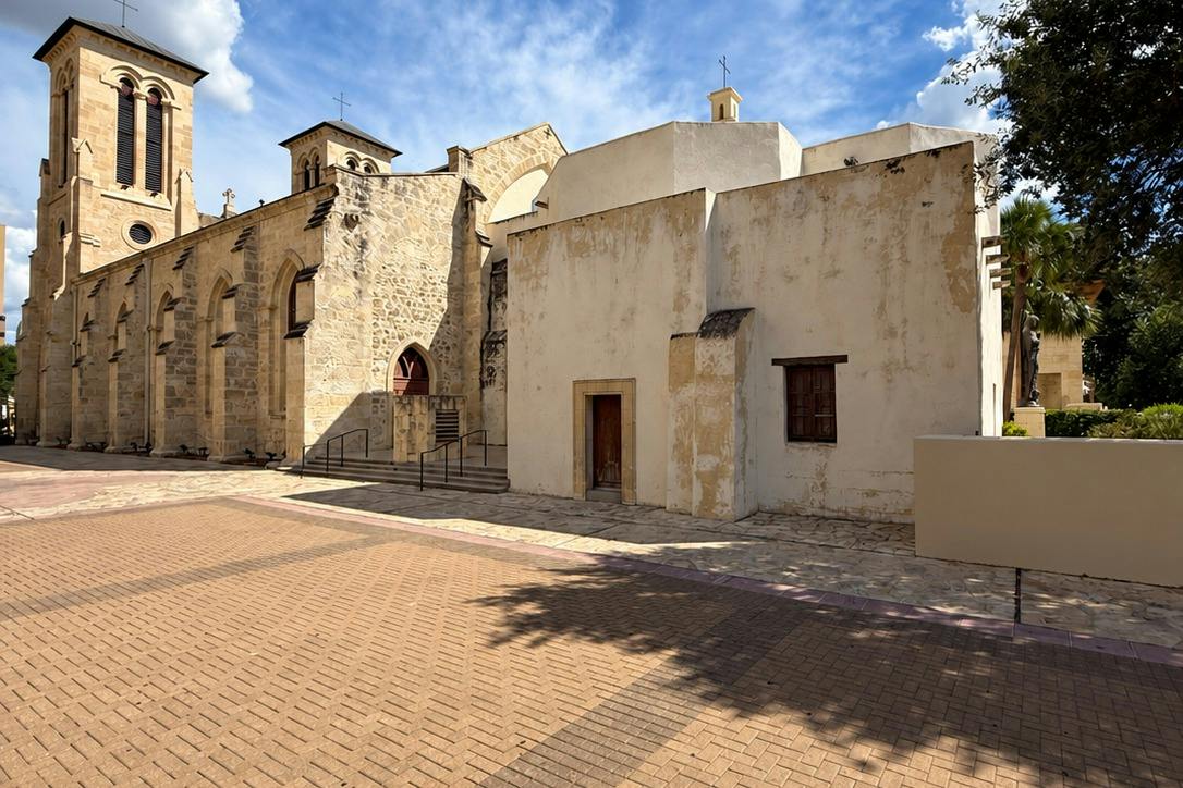 Historic stone and stucco buildings under a blue sky with a paved courtyard in the foreground.