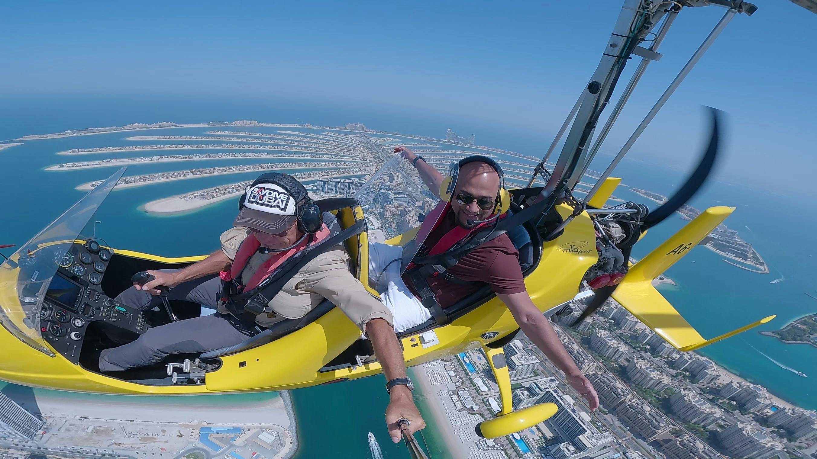 Duas pessoas a voar num girocóptero amarelo sobre uma paisagem urbana costeira com o céu azul e o oceano em fundo.