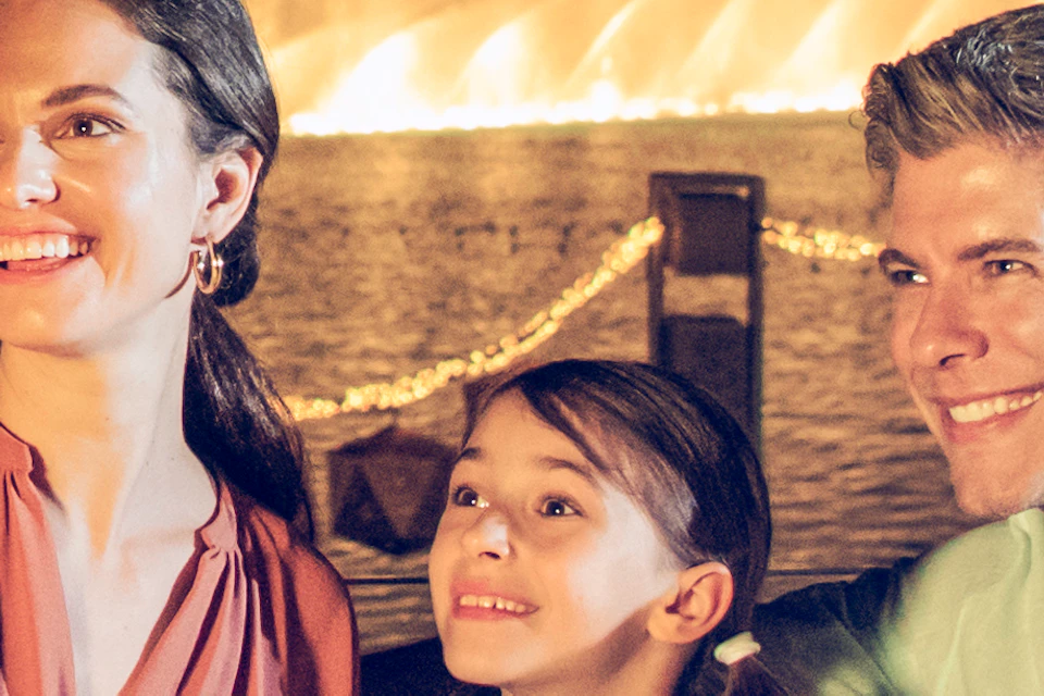 A family of three takes a selfie in front of a night-lit cityscape and a bright, illuminated fountain show.