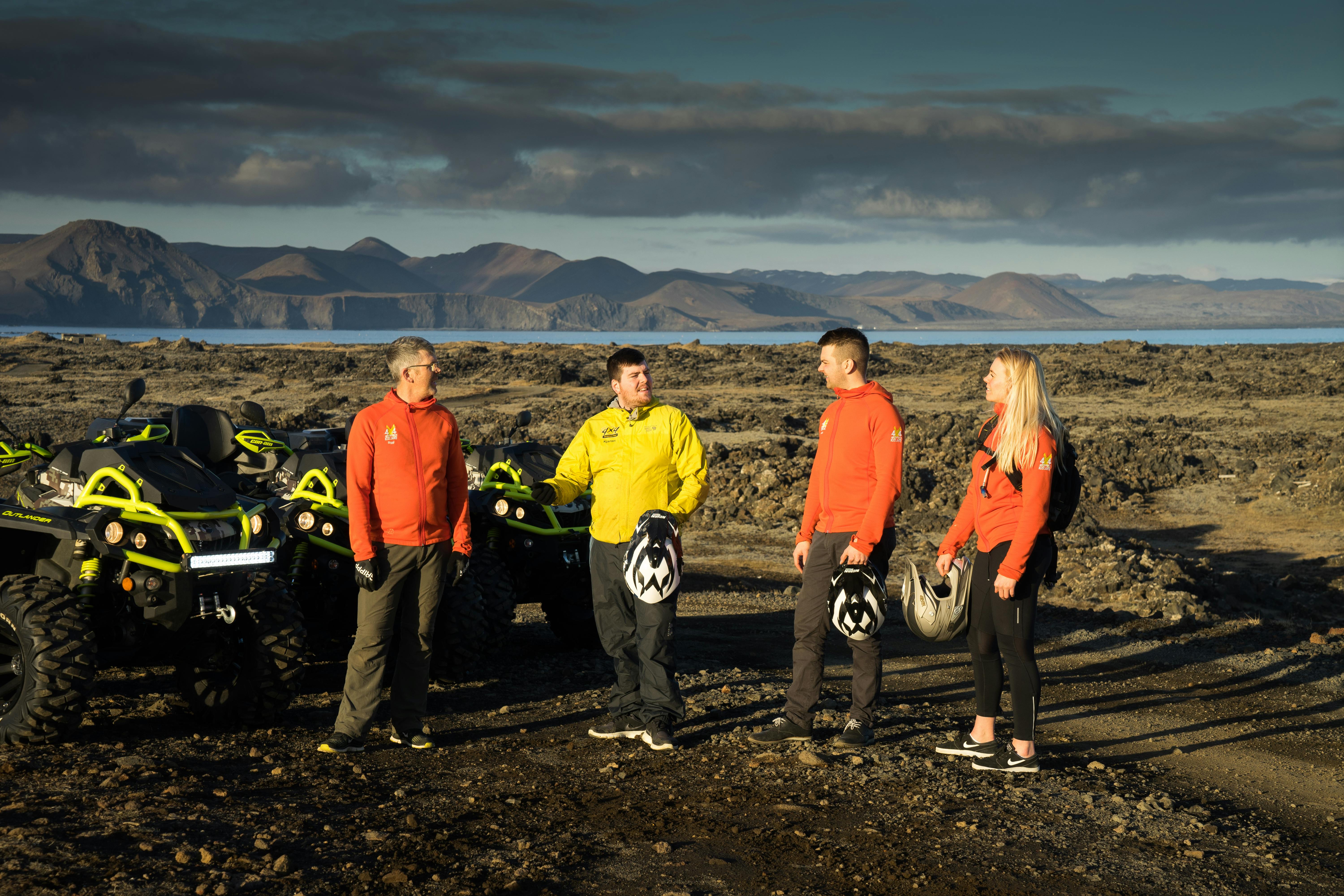 Quatre personnes vêtues de vestes vibrantes se tiennent debout, casquées, près de véhicules tout-terrain sur un terrain rocailleux, avec des montagnes et de l'eau en arrière-plan.
