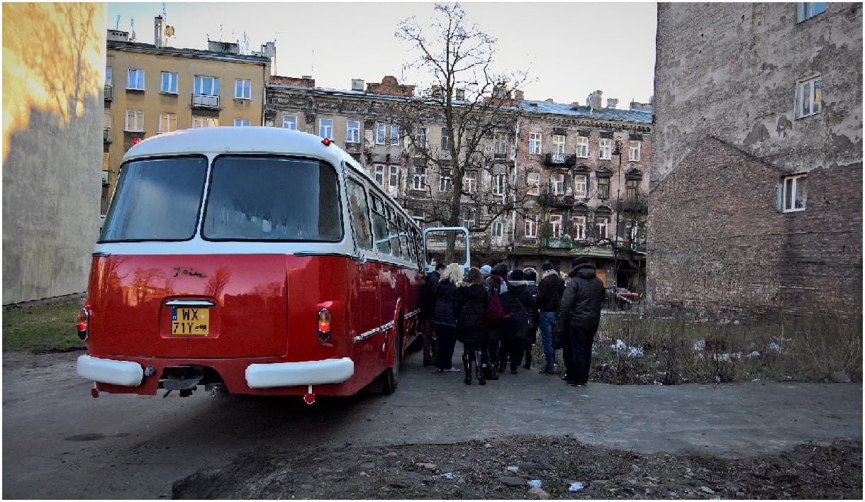 古い複数階建ての建物の前にある、赤と白のバスの横の歩道に、人々のグループが立っています。