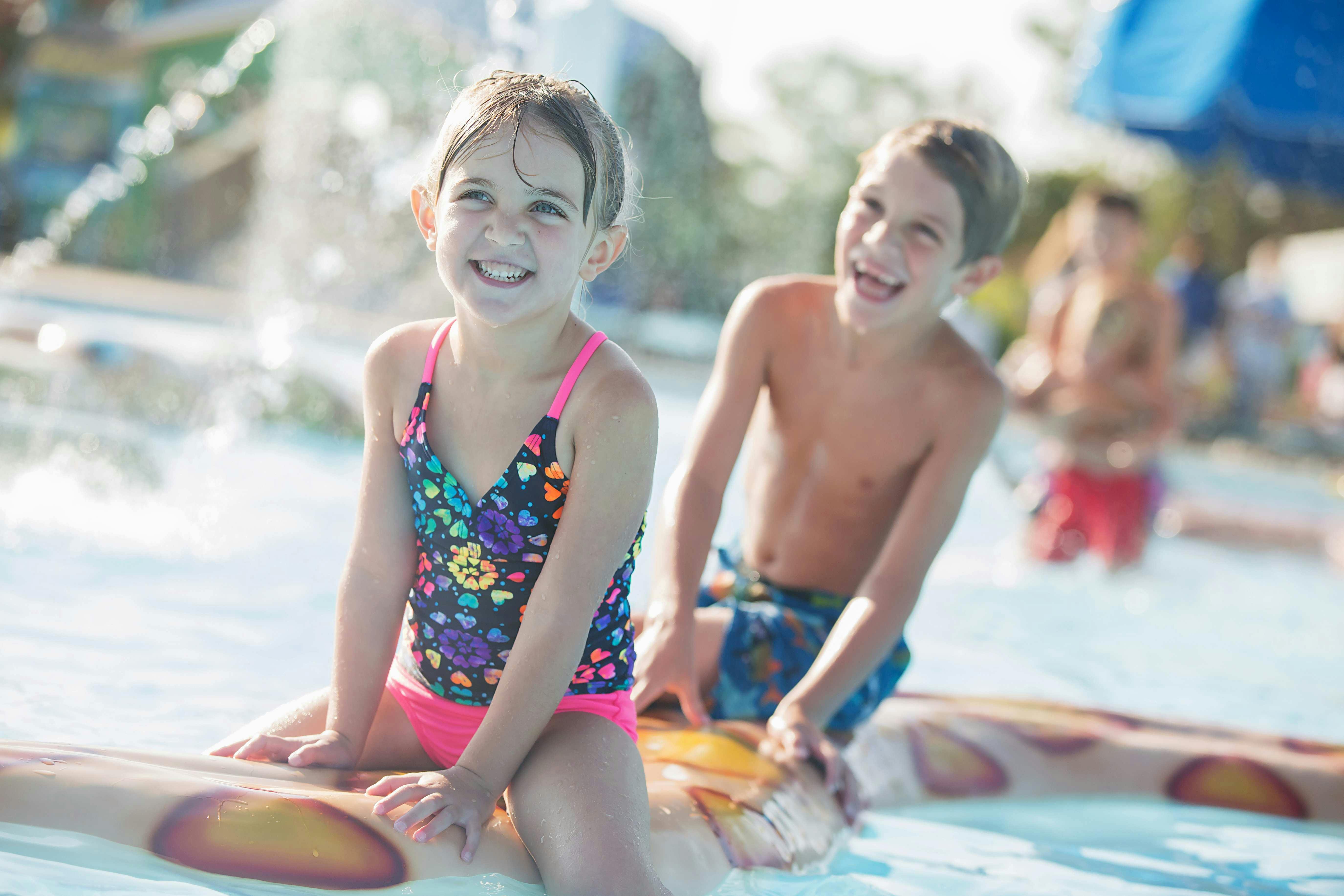Two children in swimsuits smile and play on an inflatable toy in a shallow pool with water splashing in the background.