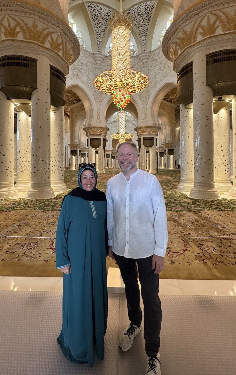 Two people standing inside an ornate hall with large columns and chandeliers. The man is wearing a white shirt; the woman wears a teal dress and headscarf.