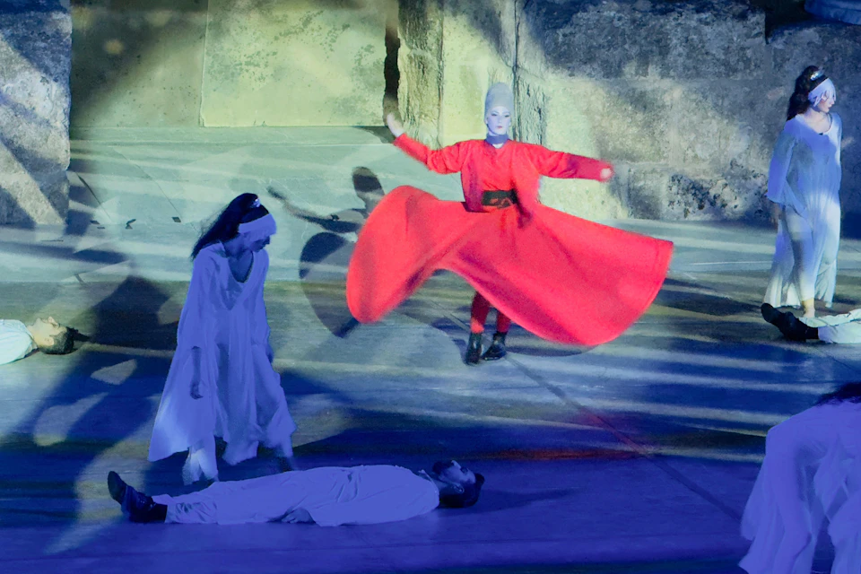 A stage performance featuring a person in a vibrant red outfit spinning, another person standing in white, and one lying down.