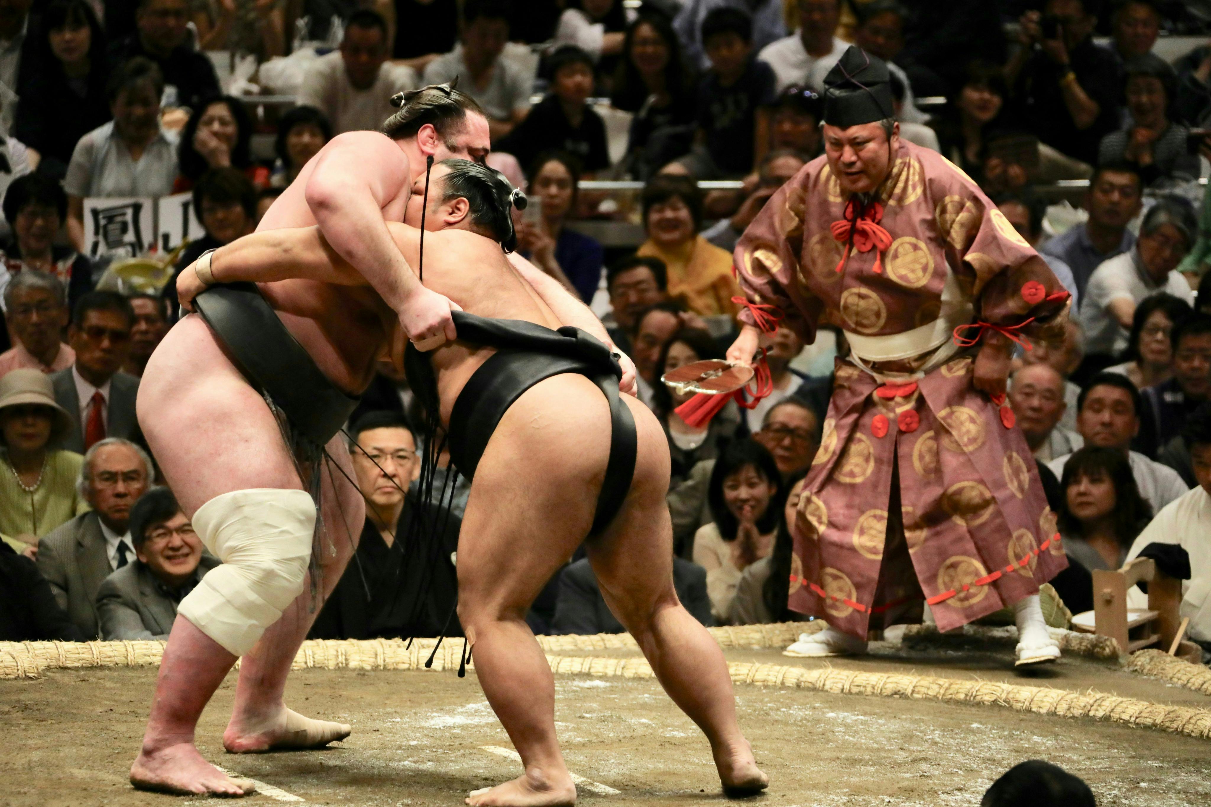 Two sumo wrestlers grappling in a ring during a match, with an official in ceremonial attire observing closely.