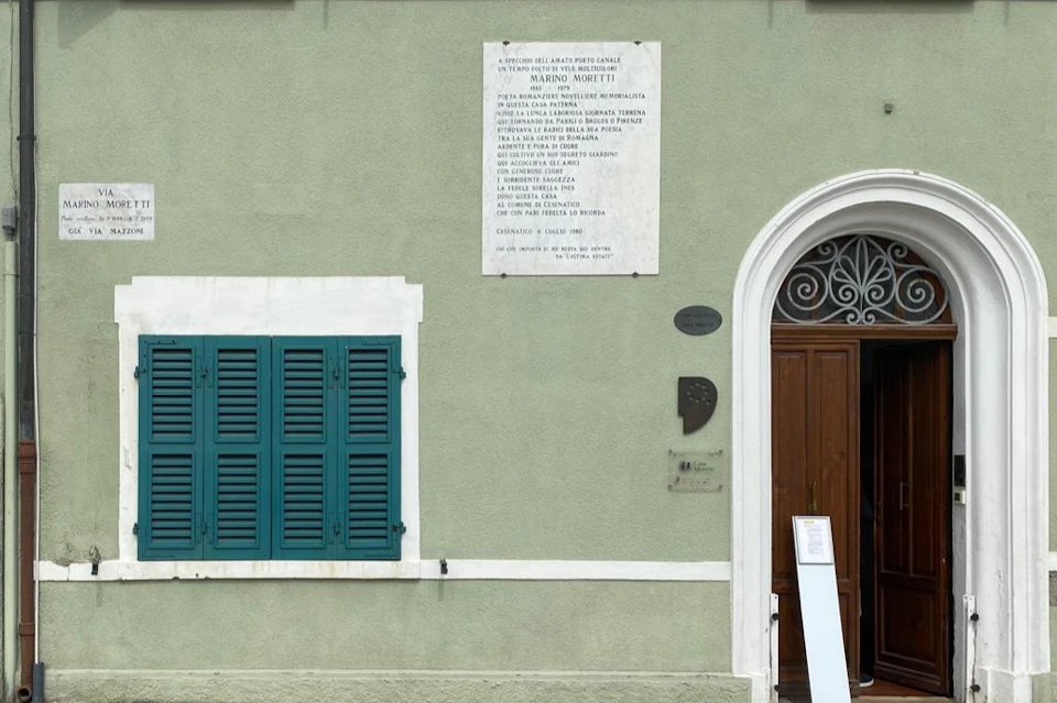 Green wall with a plaque, arched wooden door, and teal shutters. Text plaques are above and beside the door.