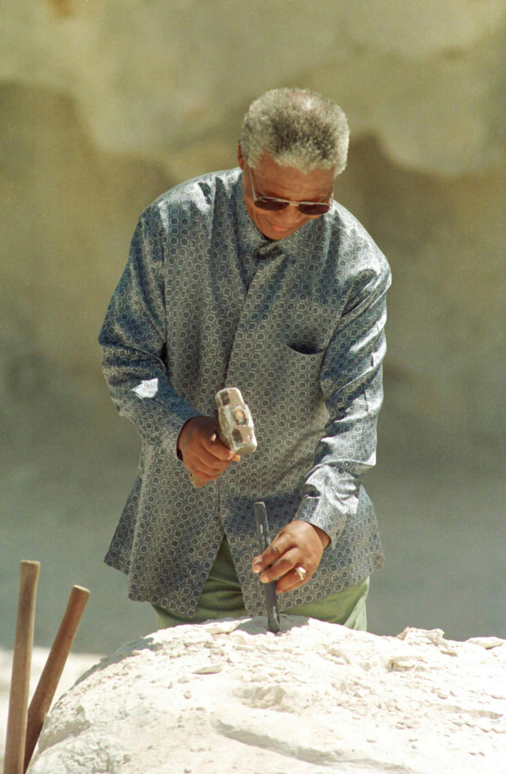 ぼやけた砂の背景をした柄のシャツを着て、短い灰色の髪とロックハンマーを持ったサングラスを持った人。