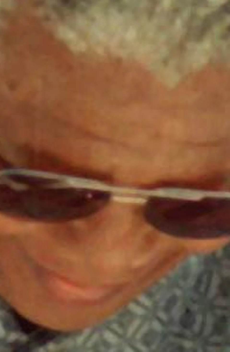Person with short gray hair and sunglasses holding a rock hammer, wearing a patterned shirt, with a blurred sandy background.