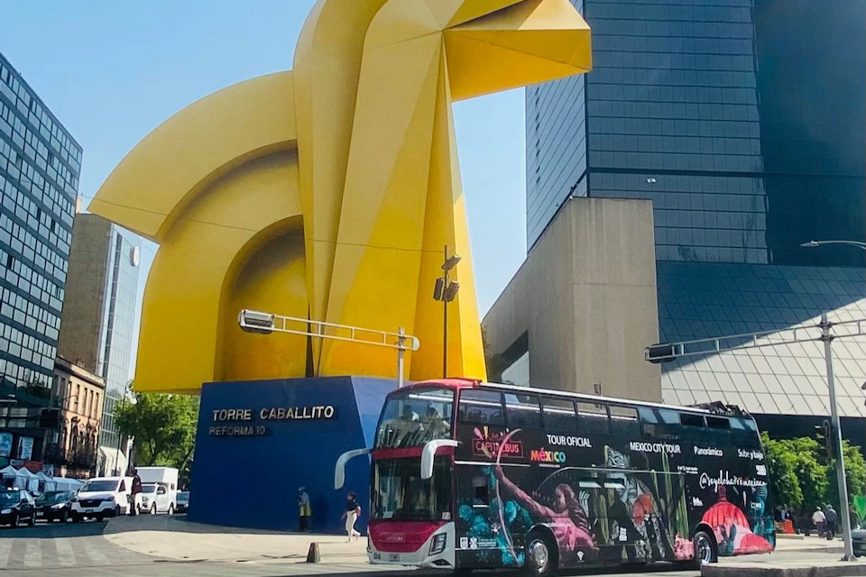 A double-decker tour bus in front of the large yellow "Torre Caballito" sculpture, with buildings in the background.