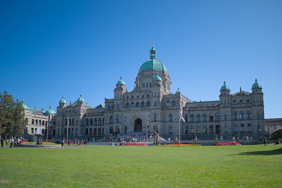 A large, historic building with green domes and a symmetrical facade. People are gathered on the steps, and a green lawn is in the foreground.