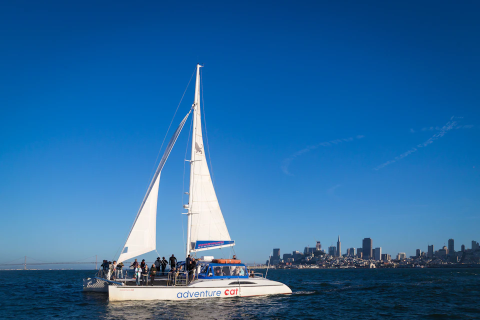 A sailboat labeled "Adventure Cat" with people on board on a sunny day, with a city skyline visible in the background.