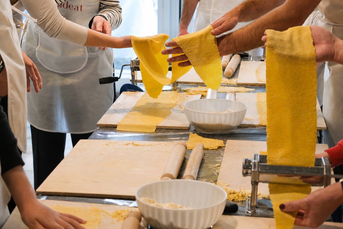 People making pasta dough at a workshop, with rolling pins, pasta sheets, and bowls on the table.