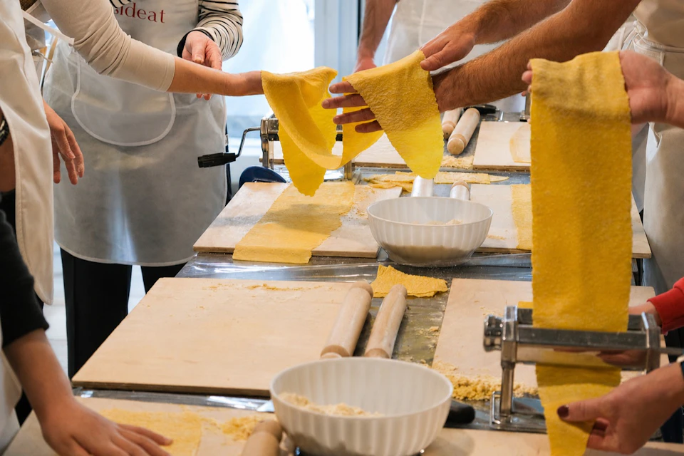 People making pasta dough at a workshop, with rolling pins, pasta sheets, and bowls on the table.