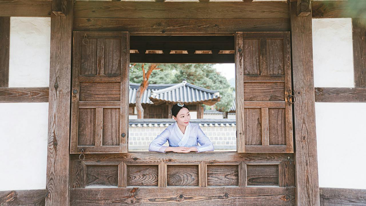 A woman in traditional Korean attire leans on a wooden windowsill, with a traditional Korean building visible in the background.