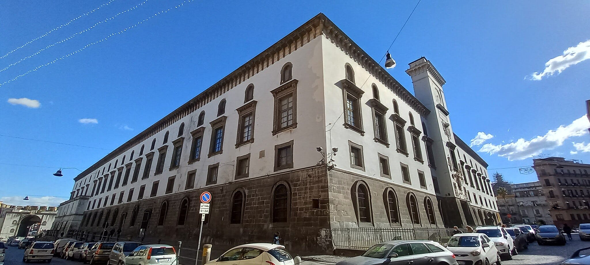 Un grande edificio storico in una giornata di sole, con finestre ad arco e numerose auto parcheggiate lungo la strada. Sono visibili alcuni pedoni.