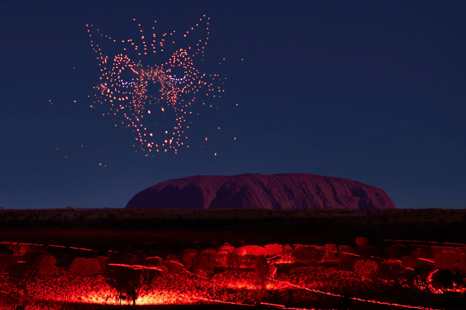 A night scene with a formation of lights in the shape of a fox's face above a landscape with red-lit foliage and a large hill.