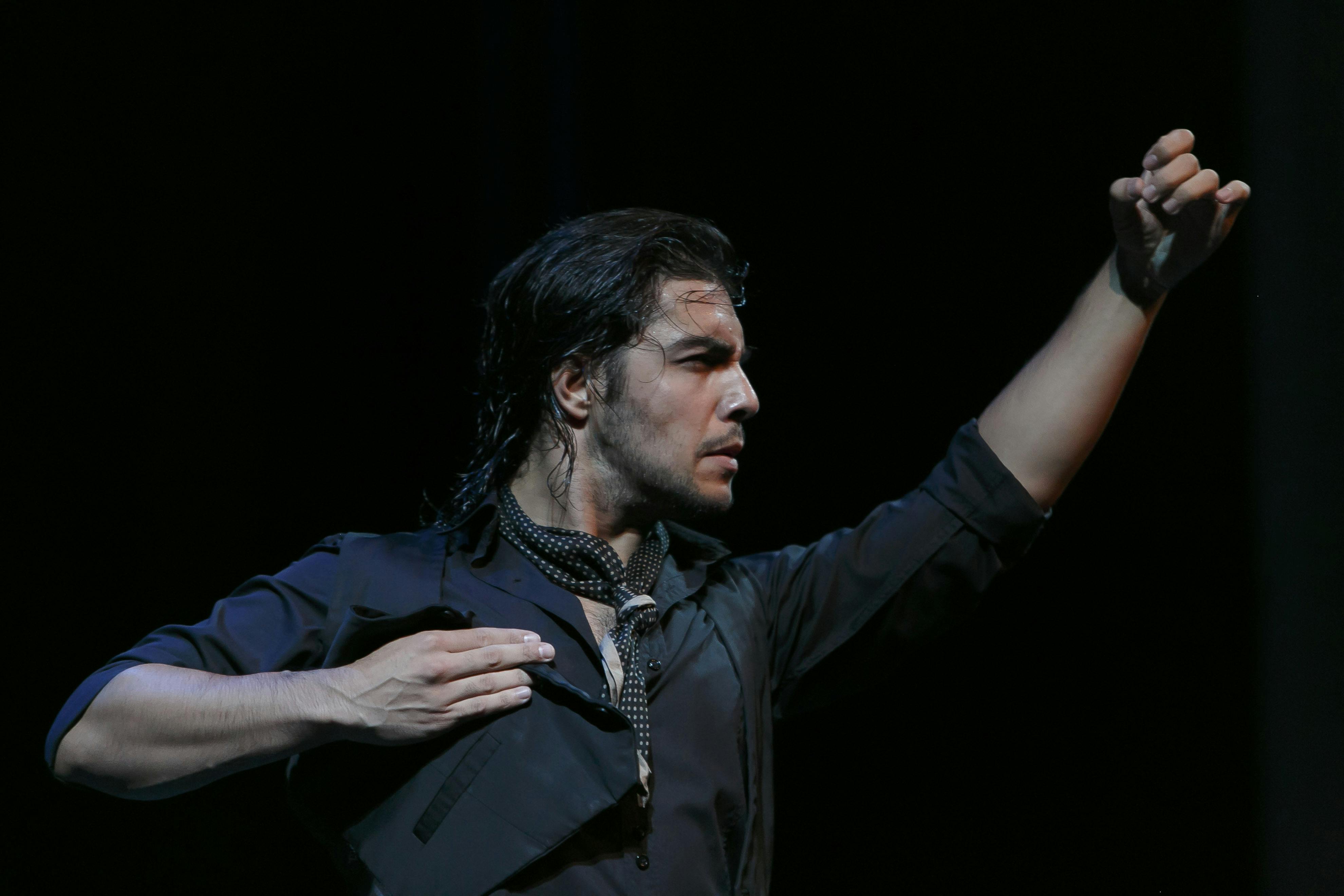 검은색 옷을 입고 젖은 머리카락과 무늬가 있는 스카프를 두른 남성이 어두운 배경에 한쪽 팔을 올린 채 극적인 포즈를 취하고 있습니다.