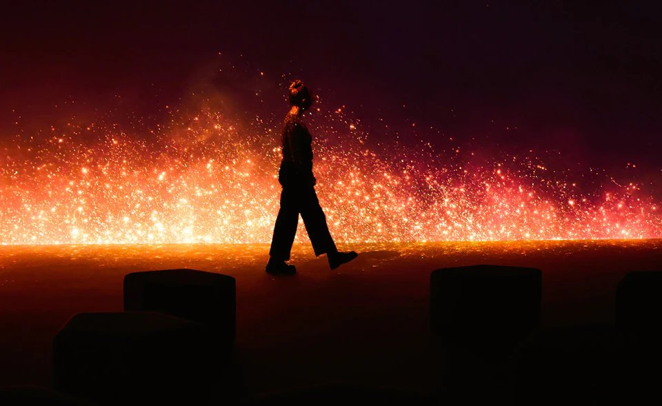 Silhouette of a person walking in front of a glowing, fiery background with vibrant sparks and a dark foreground.