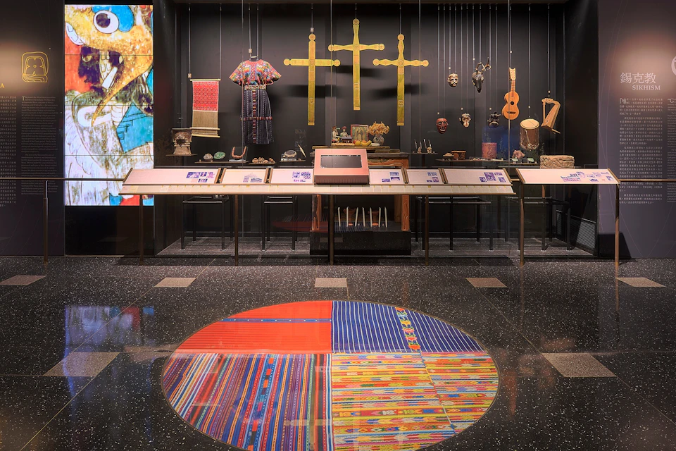 A museum exhibit with various artifacts including crosses, textiles, masks, and musical instruments displayed.