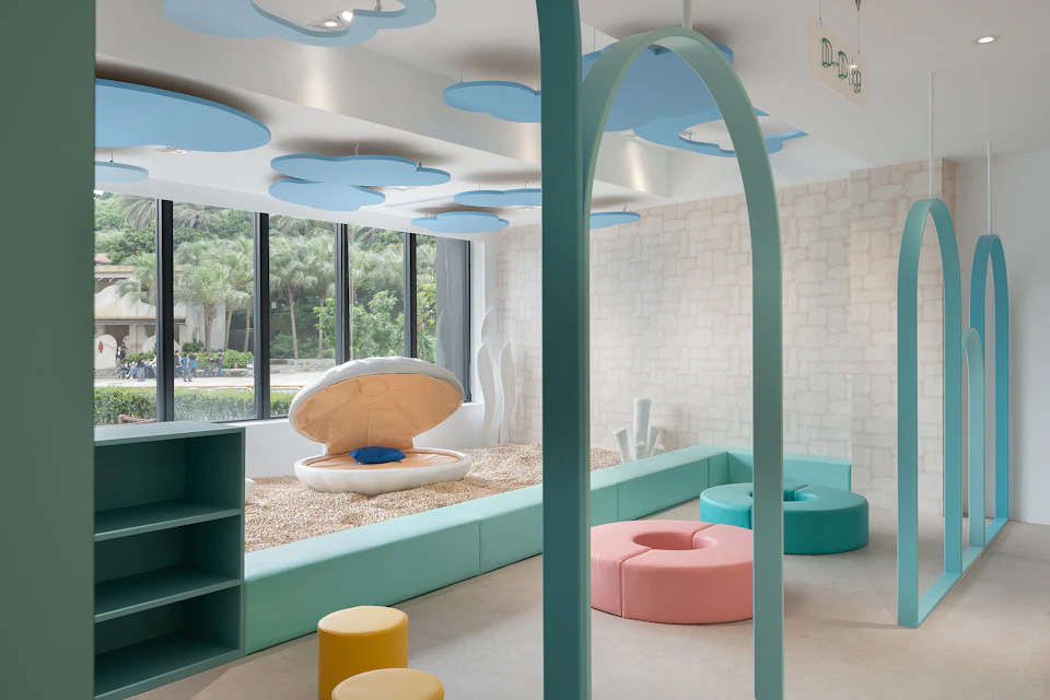 A playroom with pastel-colored furniture, a shell-shaped seat, ceiling decorations, and large windows overlooking greenery.