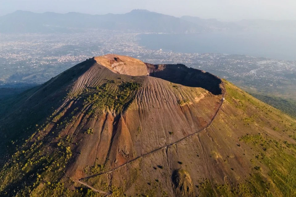 Aerial view of a large volcanic crater with a winding path on a mountain, overlooking a distant city and coastline.