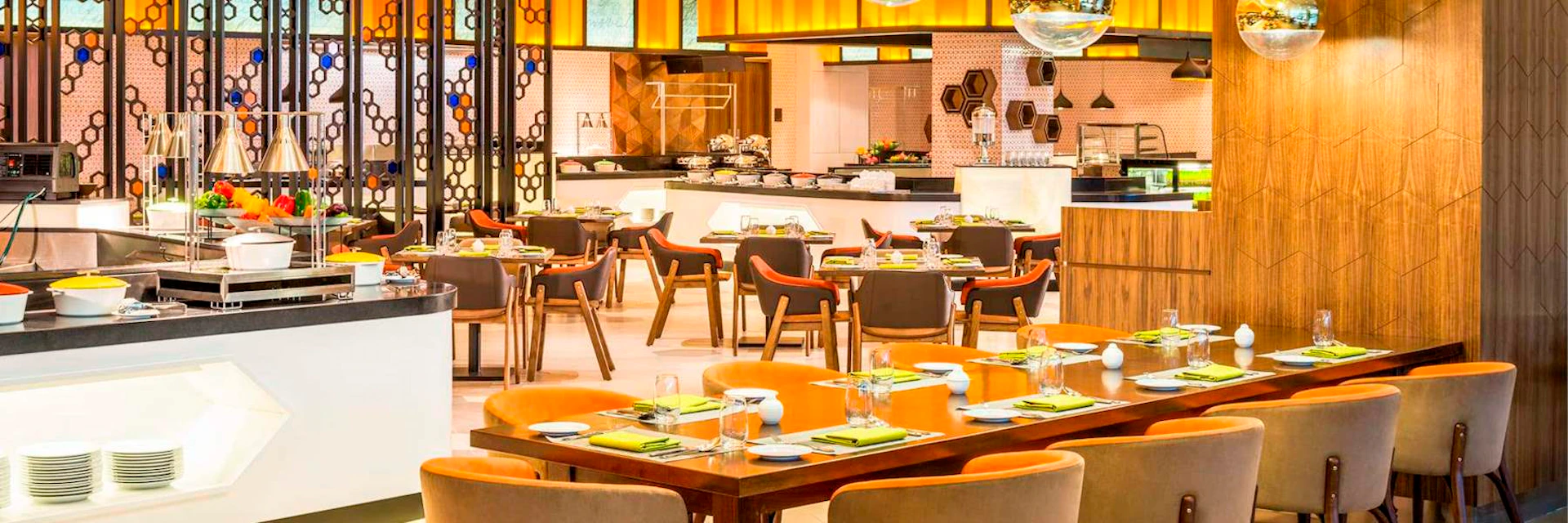 A modern dining area with neatly set tables, orange chairs, and a buffet section in the background.
