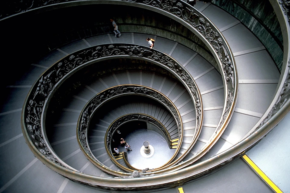 A spiraling staircase with intricate railings and people walking on it, viewed from above, leads to a circular atrium.