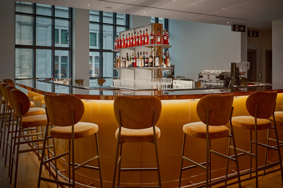 A modern bar with a curved counter, upholstered stools, and shelves displaying various liquor bottles under soft lighting.