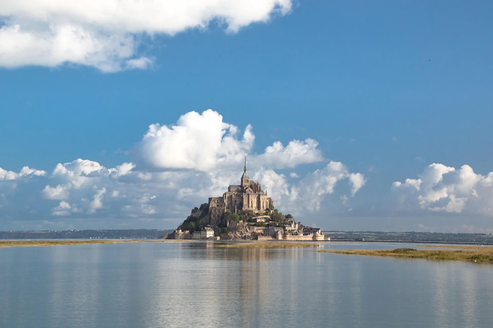 A historic island abbey sits on a mount surrounded by water, with a cloudy blue sky in the background.