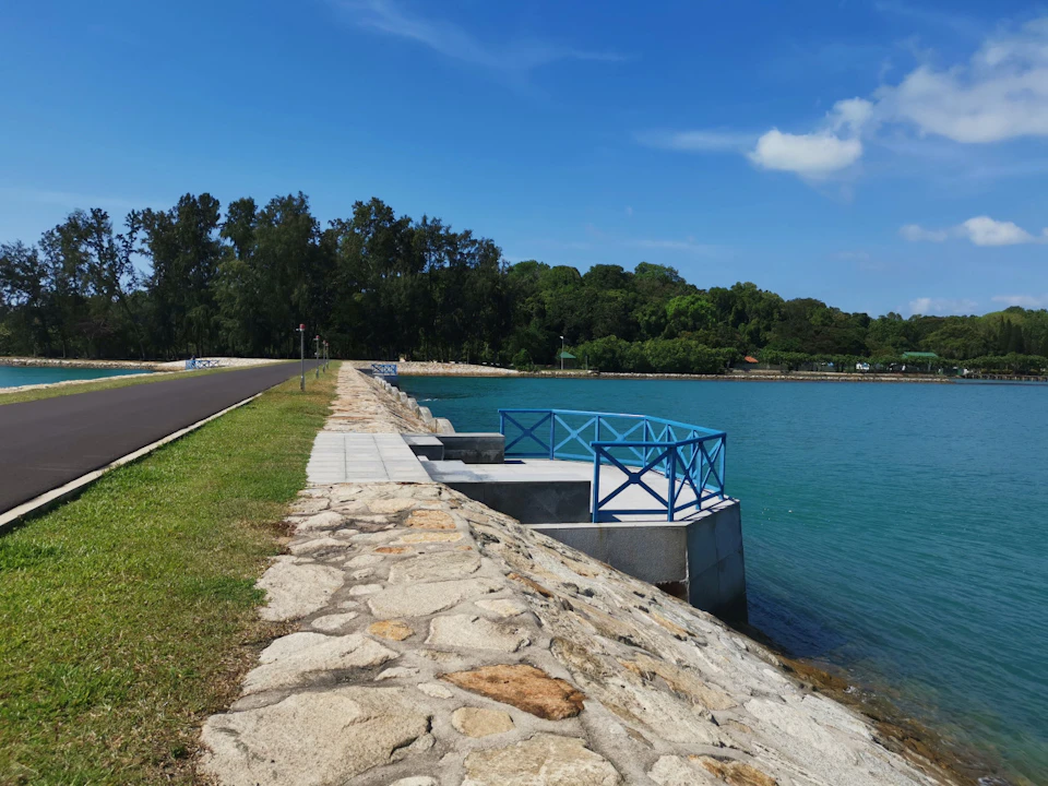 A stone walkway leads to a blue railing overlooking a calm body of water, with a grassy area and trees in the background.