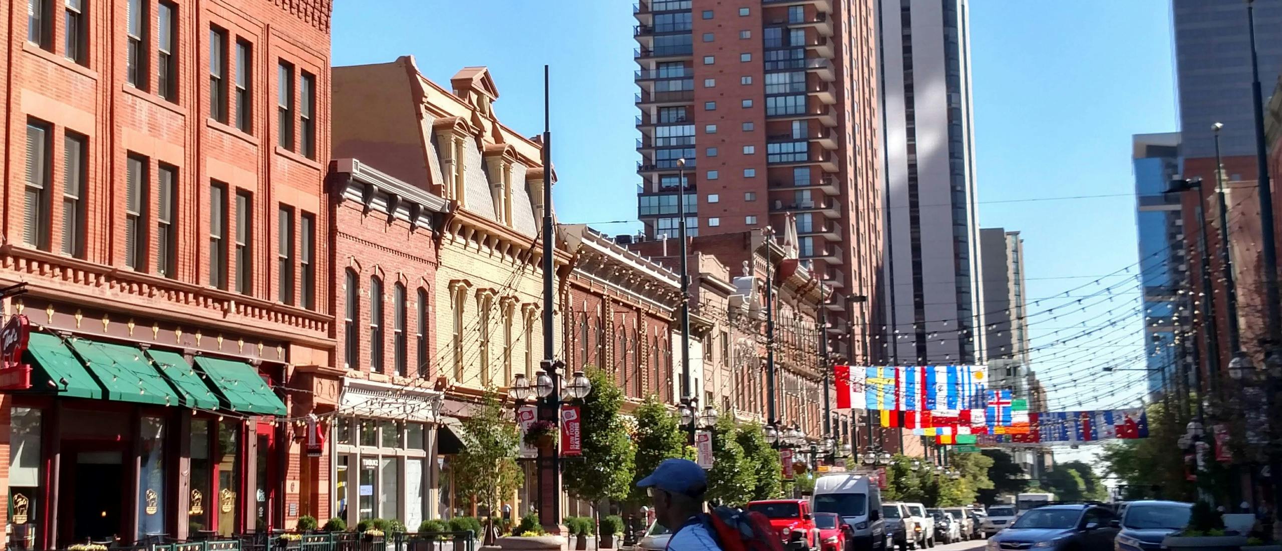 Une rue de la ville avec des bâtiments historiques en briques, des arbres, des lampadaires suspendus, un piéton et, en arrière-plan, des gratte-ciel modernes.