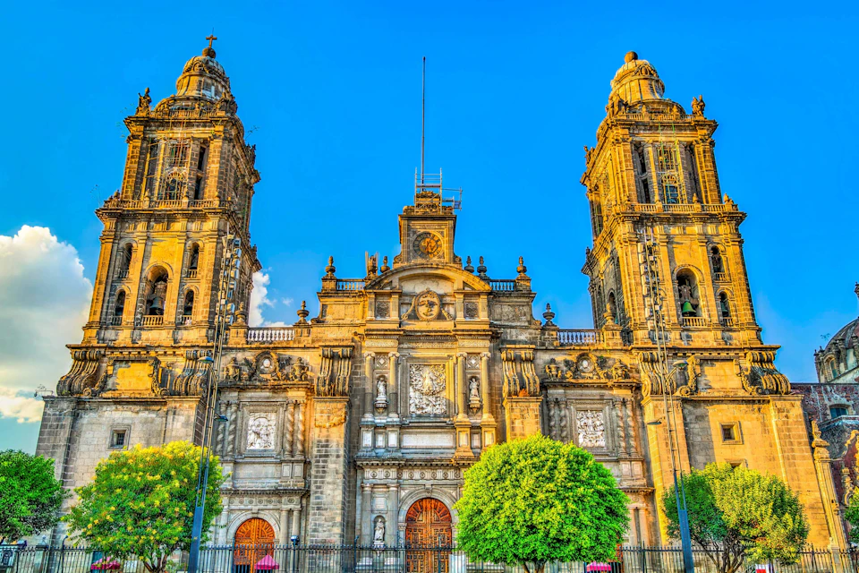 A grand, historic cathedral with two tall, ornate towers and detailed stone carvings under a clear blue sky, flanked by lush green trees.